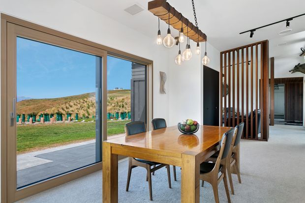 Bright dining area with a solid timber dining table and matching chairs, set beside large timber-framed sliding glass doors opening to a landscaped exterior. A distinctive suspended timber beam with multiple pendant bulbs hangs above the table, while a vertical timber slat divider separates the dining space from an adjacent interior area. The room has white walls, a pale carpeted floor, and warm natural timber detailing.