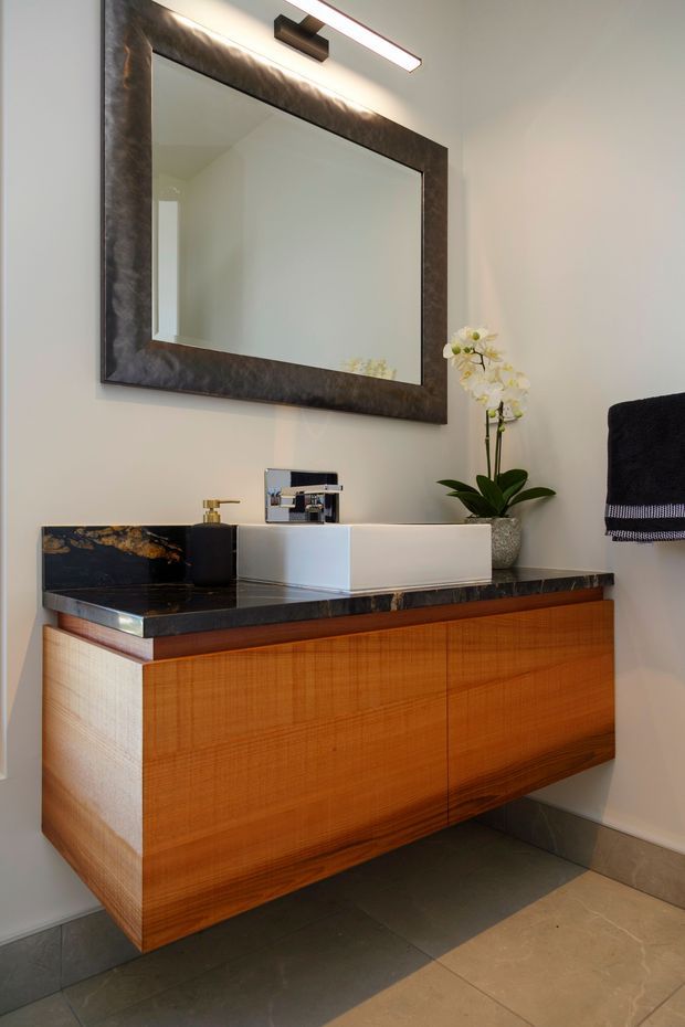 A refined contemporary bathroom vanity featuring a floating warm timber cabinet, dark stone benchtop with integrated raised white basin, chrome wall-mounted tapware, and a large dark-framed mirror with integrated lighting. Neutral walls, dark floor tiles, a black towel, and a small orchid complete the elegant composition.