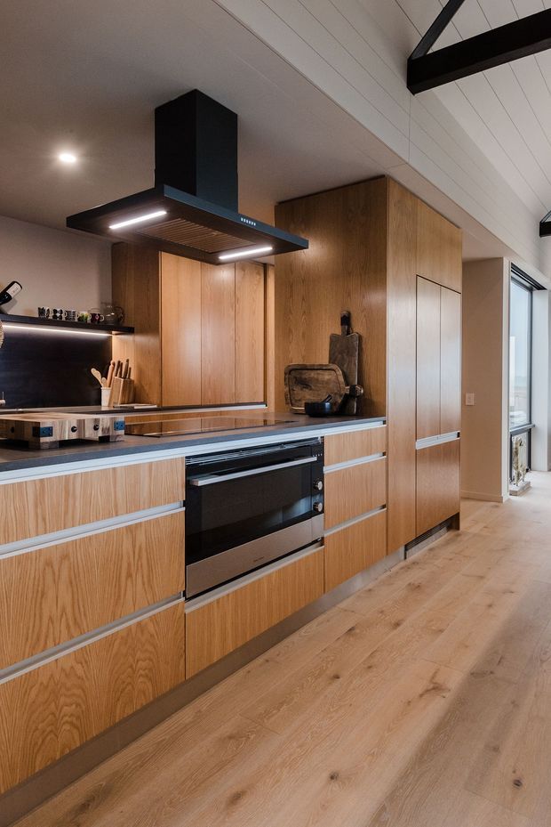 Contemporary kitchen with extensive warm timber cabinetry, integrated black appliances, a dark benchtop, and a sculptural black rangehood. The space features a timber floor, white panelled walls, open shelving with under-shelf lighting, and exposed dark structural beams near the high ceiling.