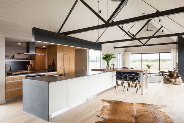 A spacious open-plan kitchen and living area with a long white island, dark benchtop, integrated sink, timber cabinetry, and bar seating. The room features high white-lined ceilings with exposed black steel trusses, large windows, pale timber flooring, and a relaxed dining and lounge zone beyond the kitchen.