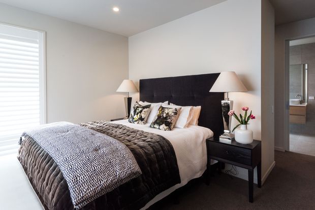 A contemporary bedroom with a queen or king bed, oversized black upholstered headboard, layered white and charcoal bedding, matching bedside tables and lamps, and neutral walls. A bathroom is visible through the open doorway at right, while a large shuttered window provides natural light.