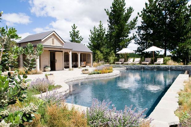 A landscaped outdoor swimming pool beside a light timber-clad pavilion or pool house with a gabled roof, covered veranda, and louvred gable detailing. Sun loungers and umbrellas sit among mature trees and ornamental planting around the pool.