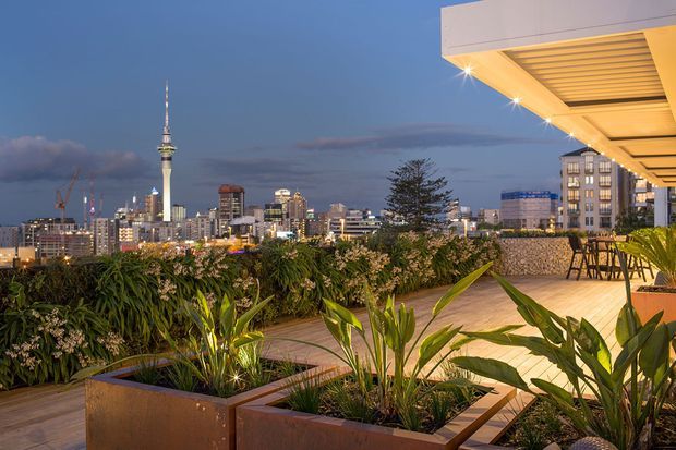Evening view from a landscaped rooftop terrace overlooking a dense city skyline, including a prominent tower. The terrace features large rust-toned planter boxes with lush tropical planting, pale tiled paving, outdoor bar seating, and a contemporary covered canopy with integrated lighting.