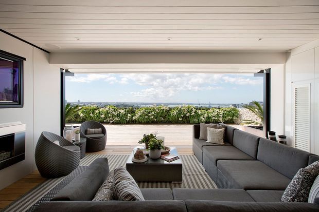 A bright, contemporary indoor-outdoor living room opening onto a timber deck and planted privacy screen. The room features a large grey sectional sofa, matching lounge chairs, a low dark coffee table, striped rug, fireplace, white walls, and a horizontal timber-lined ceiling. Wide sliding or folding glazing frames an expansive coastal or urban outlook.