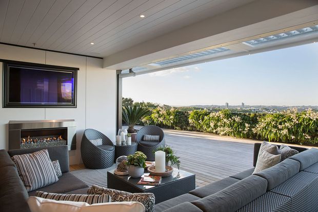A contemporary covered outdoor lounge opening onto a broad timber deck, with a large sectional sofa, armchairs, coffee table, built-in fireplace, television, and expansive views over a leafy suburban landscape. The space features a white panelled ceiling, slim structural framing, and a strong indoor-outdoor connection.