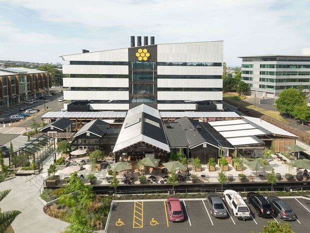 Aerial view of a contemporary mixed-use commercial complex in an urban setting. The main building has a broad white facade with horizontal dark glazing bands, a central glazed entry tower, rooftop ventilation stacks, and a yellow geometric logo. In front is a lower, dark-roofed pavilion or hospitality area with landscaped terraces, outdoor seating, pedestrian paths, and an accessible parking area.