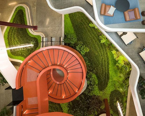 Overhead view of a contemporary interior atrium featuring a dramatic red spiral staircase, curving white balustrades, lush indoor planting, and organic-shaped circulation spaces. A blue seating area is visible at the upper right, while linear integrated lighting and grey flooring reinforce the modern design.