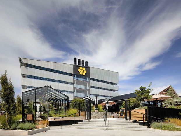 A contemporary multi-storey commercial building with a white and grey horizontally banded facade, extensive ribbon glazing, rooftop vents, and a prominent yellow hexagonal logo. The entrance forecourt includes broad concrete steps, black metal handrails, landscaped planters, a glass-framed outdoor structure, and adjacent low-rise structures with dark roofs.
