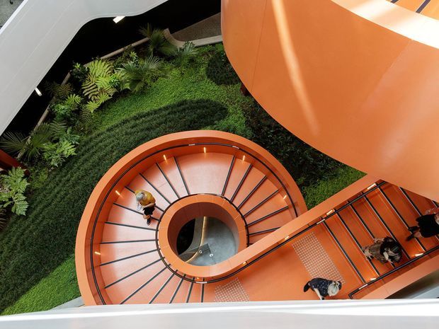 A dramatic overhead view of a sculptural orange spiral staircase winding around a central opening, with dark metal balustrades and patterned steps. Lush planted greenery fills the atrium below, while sweeping white and orange architectural forms frame the scene. Several people are visible using the staircase.