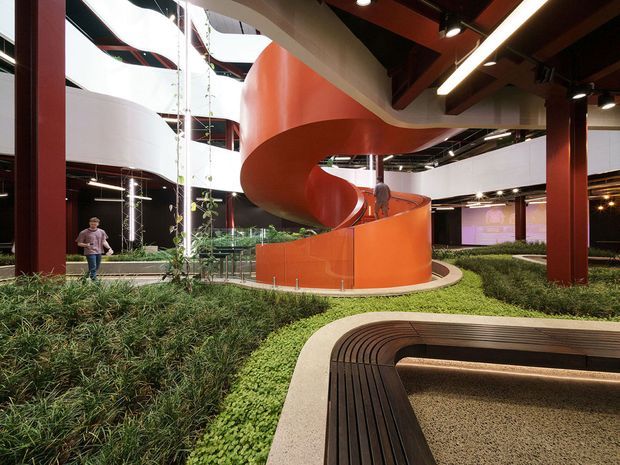 Interior view of a large contemporary atrium with a dramatic orange sculptural spiral staircase or ramp, white curved balcony edges, exposed dark timber beams, red structural columns, linear lighting, and integrated indoor landscaping. Curved built-in seating or edging surrounds a planted garden area in the foreground.