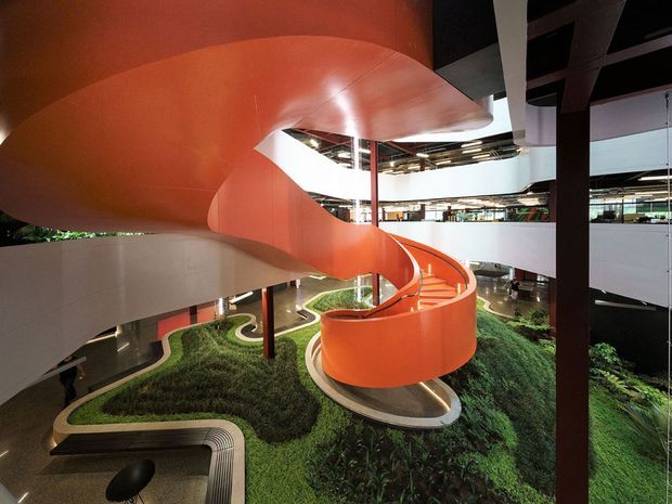 Interior architectural view of a large contemporary multi-level atrium with a striking sculptural orange spiral staircase and sweeping curved ceiling or soffit. White balcony edges, dark structural columns, exposed services, and landscaped green planting define the open central void.