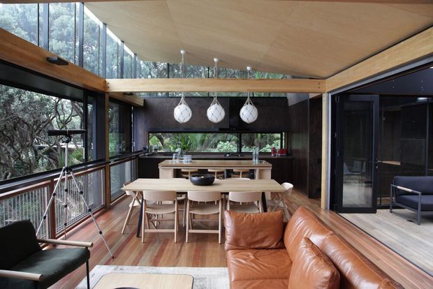 Open-plan dining and kitchen area connected to a living space, with extensive floor-to-ceiling glazing overlooking trees. The room features a timber-lined angled ceiling, warm timber flooring and joinery, dark walls and cabinetry, a long dining table, pendant lights, and a tan leather sofa.