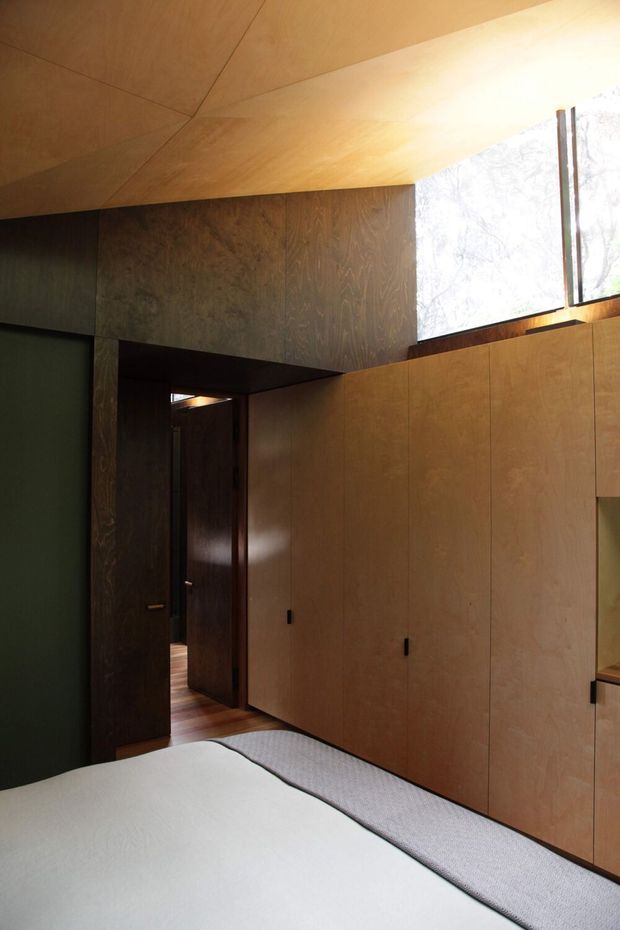 A compact, dark-toned bedroom with extensive warm timber wall and ceiling panelling, integrated full-height wardrobe cabinetry, and a partially visible bed in the foreground. A high window brings in filtered natural light, while a doorway leads to an adjacent area. The interior has a restrained, highly crafted timber-lined character.