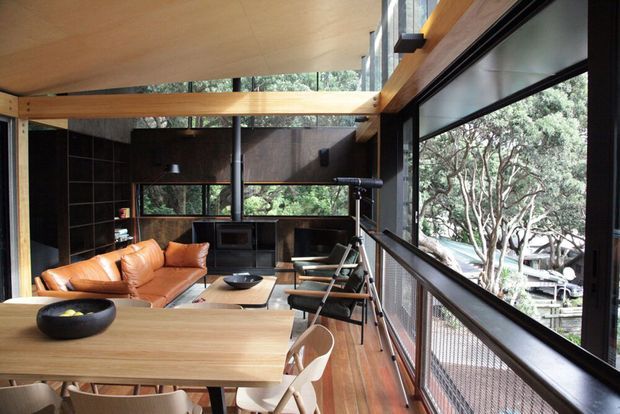 A warm, open-plan living and dining space with expansive floor-to-ceiling glazing, exposed timber beams, dark wall finishes, and a timber dining table. The room includes a tan leather sofa, fireplace or wood-burning stove, dining chairs, and a balcony-like glazed edge overlooking trees. The design combines natural timber, black metal framing, and a strong indoor-outdoor connection.