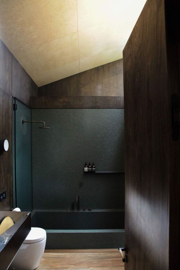 Compact dark bathroom with a bathtub and shower at the rear, wall-mounted toilet, dark textured wall tiles, timber-look wall panels and flooring, and a pale angled ceiling that creates a distinctive sloping form. Minimal black and brushed-metal fixtures contribute to a moody contemporary atmosphere.