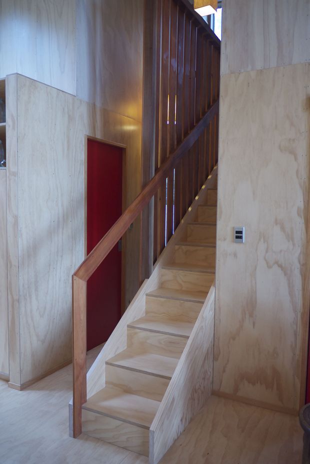 Interior view of a compact timber staircase rising through a plywood-lined space. The stair treads, surrounding walls, and balustrade are predominantly pale plywood, with a dark stained timber slatted screen and handrail forming the stair enclosure. A deep red door and small wall switch provide contrasting details.