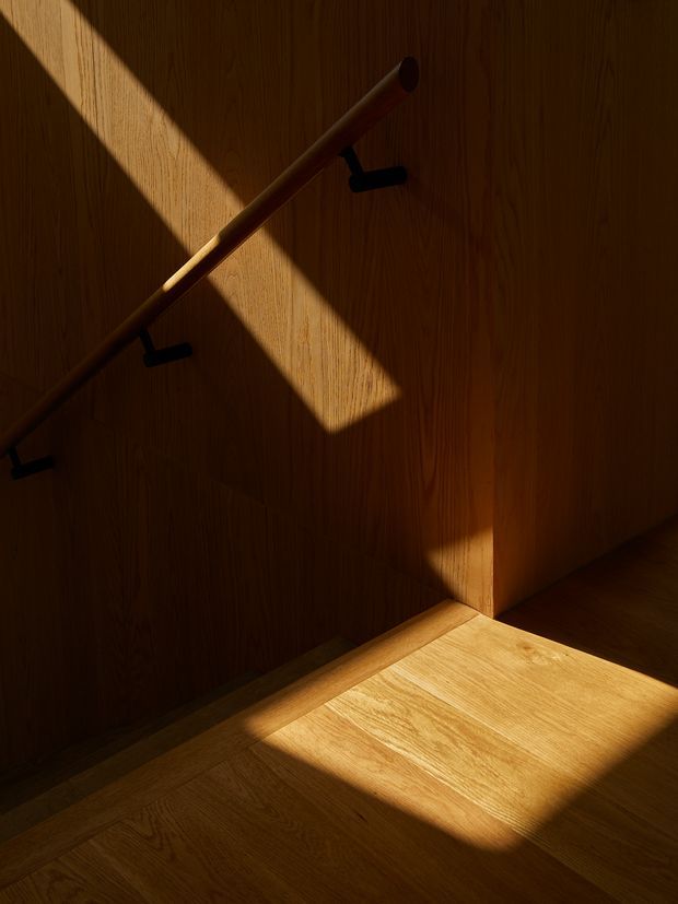 A moody, close-up architectural detail of a timber-lined stairway. Warm oak-toned wall panelling and matching timber flooring are crossed by strong diagonal sunlight, while a simple dark handrail runs along the wall. The image emphasizes natural wood grain, shadow, and geometric light.