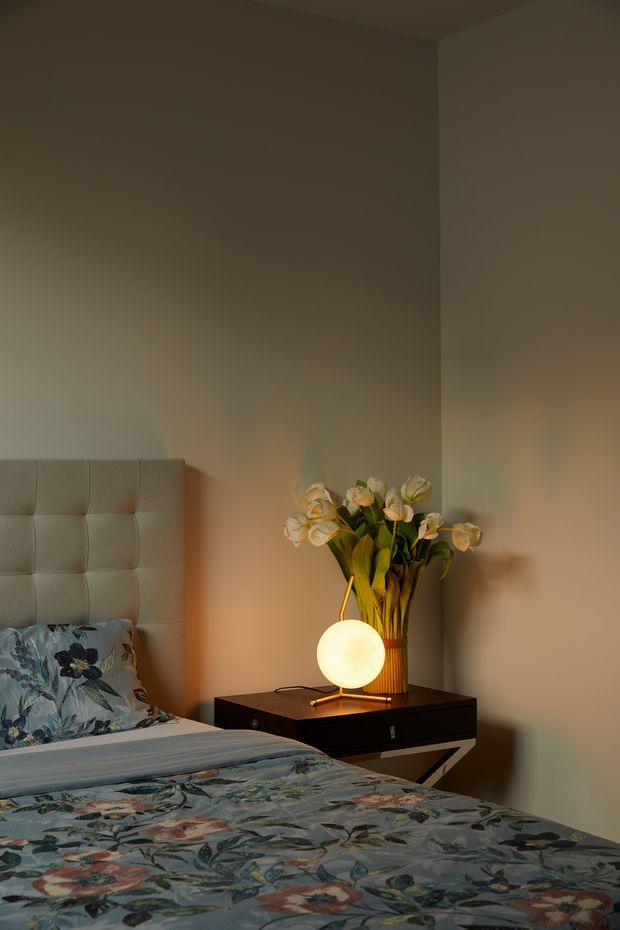 A warmly lit bedroom corner featuring a partially visible upholstered, vertically tufted headboard, a bed dressed in patterned floral bedding, and a dark floating bedside table with a spherical glowing lamp and tall vase of flowers. Neutral taupe walls and soft ambient lighting create an intimate, cozy atmosphere.