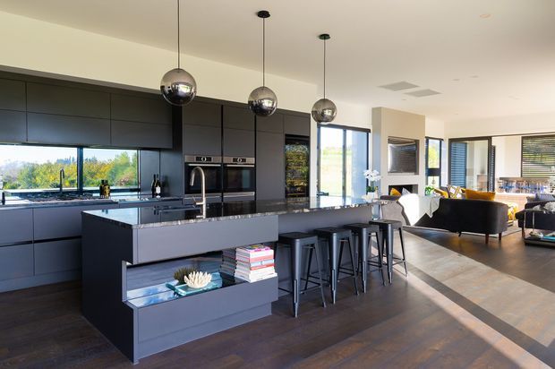 A spacious open-plan contemporary kitchen and living area. The kitchen features dark matte built-in cabinetry, a large stone island with integrated open shelving, bar seating for four, stainless appliances, and three spherical pendant lights. Timber flooring continues into the adjacent living room, which has large windows and relaxed modern furnishings.
