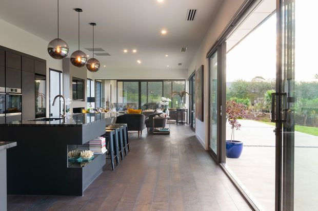 Open-plan contemporary kitchen, dining, and living area with dark integrated cabinetry, a large island with seating, timber-look flooring, pendant lights, and extensive floor-to-ceiling glazing opening onto an outdoor terrace and garden. The space is bright and connected to the outdoors, with a lounge and dining area visible beyond the kitchen.