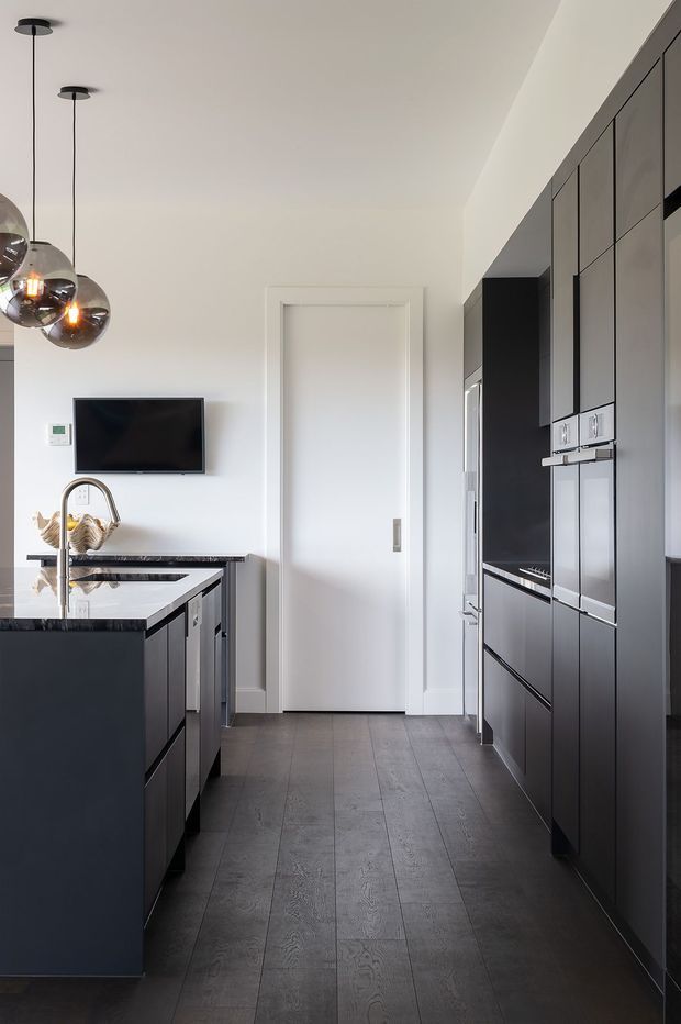 A sleek contemporary galley kitchen with dark full-height cabinetry, integrated appliances, a central island with a dark stone benchtop, and dark timber-look flooring. White walls and ceiling provide contrast, while three smoked-glass pendant lights hang above the island. A wall-mounted television and concealed door are visible at the rear.