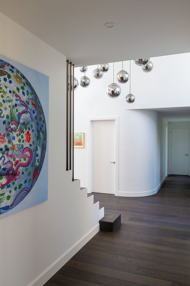Bright contemporary residential hallway and stairway with white walls, dark timber flooring, a sculptural open stair, polished metal bal pendant lights, and a large colourful botanical artwork. The corridor extends toward multiple white doors, while a curved wall adds architectural softness.