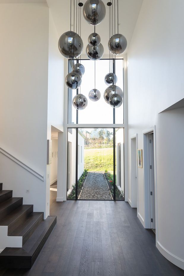A bright, contemporary residential entry hall with a double-height ceiling, dark timber staircase and flooring, white walls, and a dramatic cluster of chrome spherical pendant lights. Full-height glazing and a glazed door open toward a small landscaped courtyard, while interior doorways line the hall.