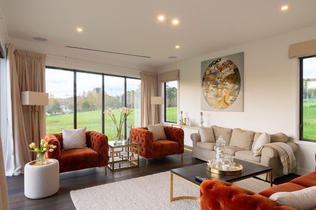 Bright, refined living room with full-height glazing overlooking a green landscape. The space features a cream sofa, rust velvet tufted armchairs and seating, a glass-topped coffee table with brass-toned frame, dark timber flooring, neutral curtains, recessed lighting, and a large decorative wall artwork.