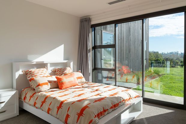 Bright modern bedroom with a white platform bed, orange-and-white patterned bedding, grey carpet, neutral walls, and full-height black-framed glazing opening to a green outdoor landscape. Grey curtains and a streamlined ceiling air vent complete the contemporary setting.