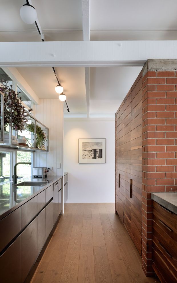 A narrow, bright kitchen with a galley-style layout. The room features warm timber flooring, dark wood-toned lower cabinetry, a stainless-steel sink and benchtop area, open shelving with plants beside large windows, and a prominent exposed red-brick fireplace or chimney structure with timber detailing. White-painted exposed ceiling beams and track lighting create a light, functional interior.