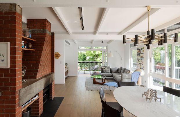 A bright open-plan living and dining area with a prominent exposed red-brick fireplace and shelving, pale timber flooring, white coffered ceiling beams, large glazed doors and windows, a grey sectional sofa, and a contemporary dining table with sculptural pendant lighting. The space opens toward a leafy outdoor balcony or garden.