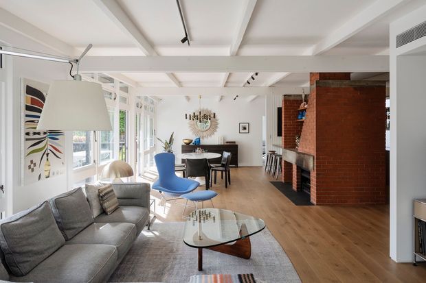 Bright open-plan living and dining area with a mid-century-inspired interior. The room features a grey sectional sofa, sculptural glass coffee table, blue lounge chair, timber dining table and chairs, white walls, extensive glazing, light timber flooring, exposed white ceiling beams, track lighting, and a substantial exposed red-brick fireplace or chimney structure. A kitchen or bar area is partially visible beyond the dining zone.
