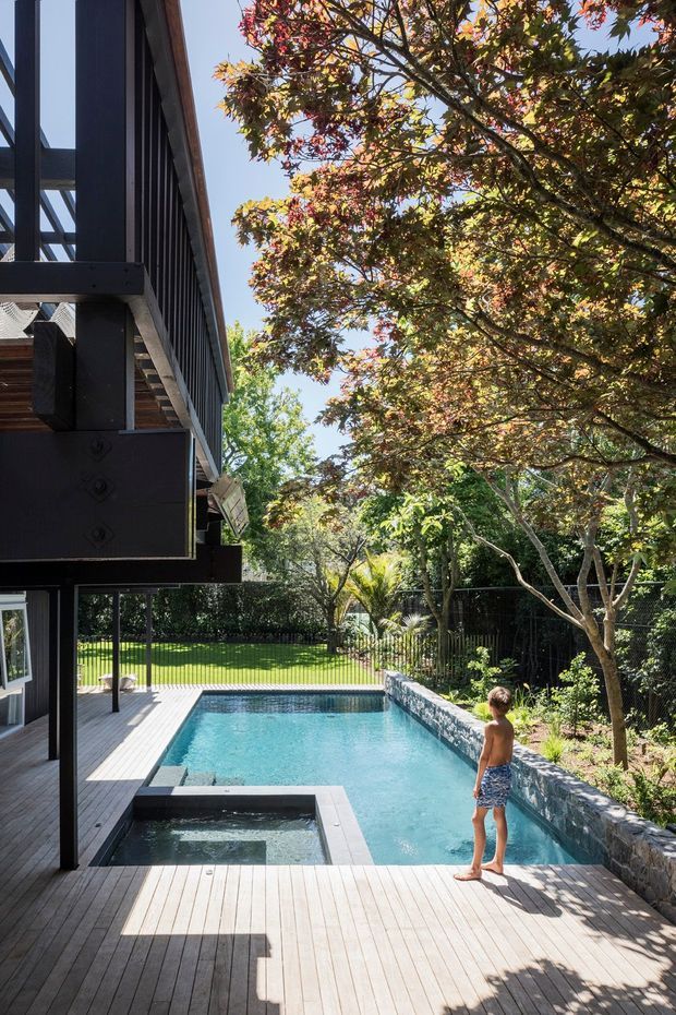 Exterior view of a contemporary, dark-clad multi-level home opening onto a timber pool deck. A long rectangular swimming pool includes a raised spa or plunge section, bordered by a textured stone retaining wall and landscaped garden with mature trees. Strong vertical facade screening, exposed structural framing and extensive glazing are visible on the left.