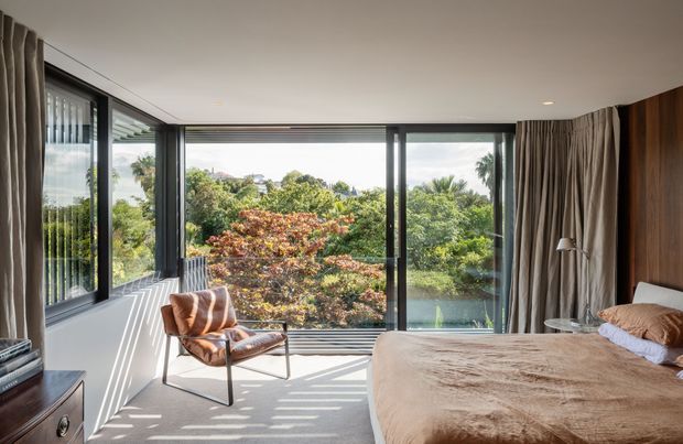 Bright contemporary bedroom with a large corner of floor-to-ceiling glazing overlooking lush greenery. The room features a low-profile bed with neutral bedding, a cognac leather lounge chair, full-height taupe curtains, dark timber wall panelling, and carpeted flooring. Strong sunlight creates patterned shadows across the floor and walls.