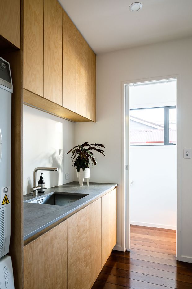 A compact, modern kitchen or kitchenette with full-height vertical-grain timber cabinetry, a dark grey stone benchtop, integrated stainless-steel sink and tap, and a white splashback. A small potted plant sits on the counter. Dark polished timber flooring leads to a bright doorway, with an adjacent utility appliance partially visible at left.
