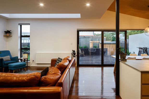 A warm, open-plan living and kitchen area with polished dark timber flooring, a large caramel leather sectional sofa, and a teal armchair. The room has white walls, recessed ceiling lighting, a horizontal clerestory-style opening, and full-height glazed sliding doors leading to a timber-decked outdoor courtyard. A kitchen island with a light benchtop is visible on the right, while a timber-lined ceiling and reflective glazing add architectural interest.
