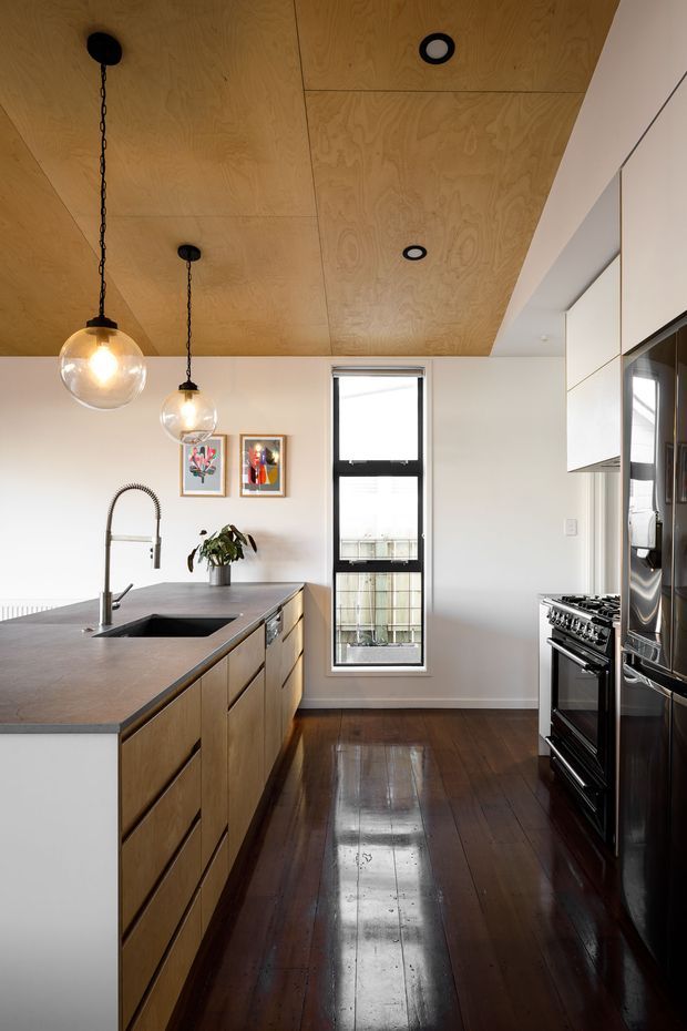 A contemporary galley kitchen with a large timber island, dark stone benchtop, undermount sink and sculptural gooseneck tap. Flat-panel timber cabinetry continues along the island, while stainless-steel cooking appliances and dark cabinetry occupy the opposite wall. The room features a warm plywood-lined ceiling with recessed downlights, polished dark timber flooring, white walls, a tall narrow window, and pendant globe lights.
