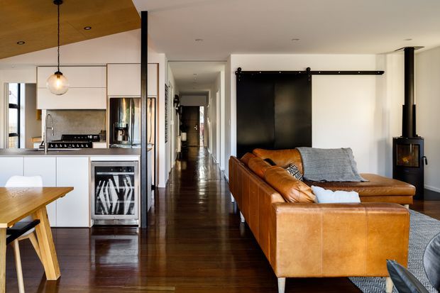 Open-plan contemporary living space combining a compact white kitchen and dining area with a leather sectional lounge. Dark polished timber flooring runs through the room and hallway, while a black sliding barn-style door and freestanding wood-burning stove add strong architectural features. The kitchen includes white fixed cabinetry, a dark benchtop, stainless-steel appliances, and a warm timber ceiling panel over the dining area.