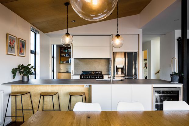 A warm, contemporary open-plan kitchen and dining area. The kitchen features a large dark benchtop island with timber detailing, white handleless cabinetry, integrated appliances, a stainless-steel oven, and a textured grey splashback behind the freestanding cooktop. Three black-framed timber bar stools sit at the island, while a timber dining table with white chairs occupies the foreground. The room has a timber-lined ceiling, white walls, black-framed windows, pendant globe lights, open shelving, and indoor plants.