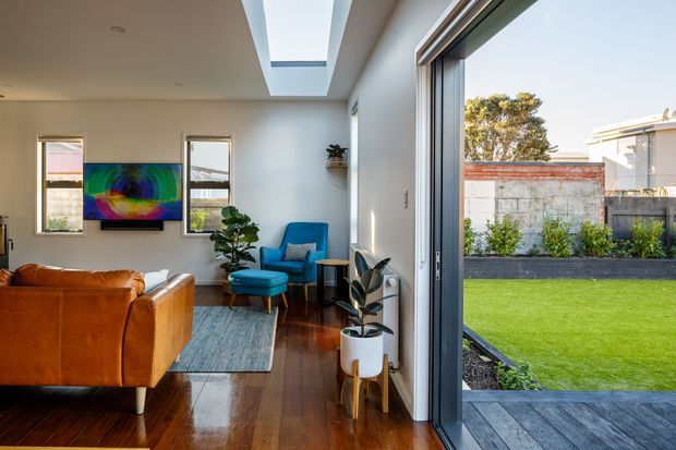 A bright contemporary living room opening directly onto a landscaped backyard through wide sliding glass doors. The room features polished dark timber flooring, a large caramel leather sofa, a blue lounge chair and ottoman, a textured area rug, potted plants, and colourful wall art. A linear skylight brings additional daylight into the space, while the exterior view shows a timber deck, lawn, raised garden bed, boundary wall, and neighbouring modern building.