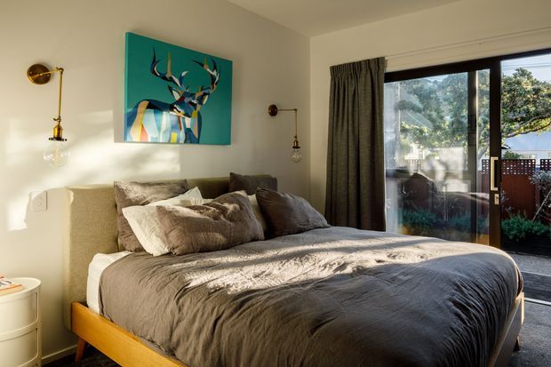 A warm, sunlit bedroom with a low-profile timber bed, layered taupe and grey bedding, and neutral upholstered elements. A colourful deer artwork provides a teal accent above the bed, while brass wall-mounted lamps flank the artwork. Full-height glazing with dark curtains opens the room visually toward a timber outdoor deck or balcony.