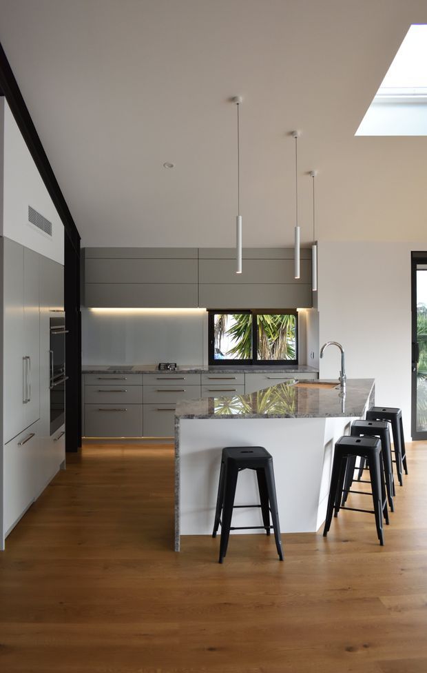 A contemporary open-plan kitchen with a large white island, waterfall stone benchtop, integrated grey cabinetry, stainless-steel appliances, and black bar stools. Warm timber flooring contrasts with the cool grey joinery. Linear pendant lights, under-cabinet lighting, a skylight, and large glazed openings create a bright, refined space.