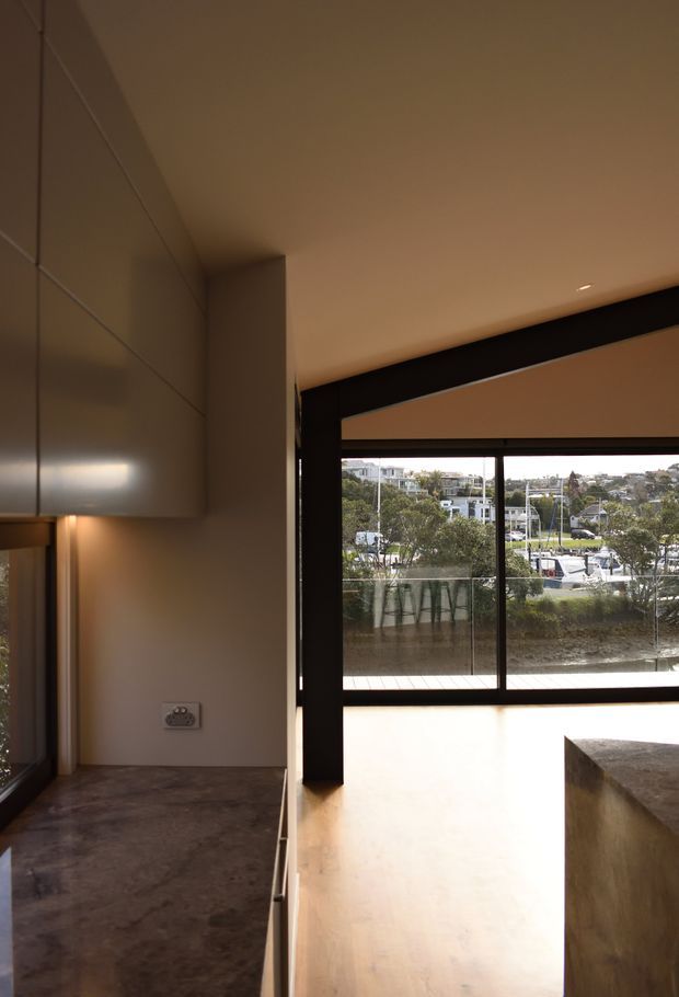 A warm-toned contemporary kitchen interior with dark stone or concrete benchtops, streamlined glossy cabinetry, and black-framed floor-to-ceiling glazing. The large window overlooks a marina or waterside urban setting, while a substantial island or counter occupies the foreground.