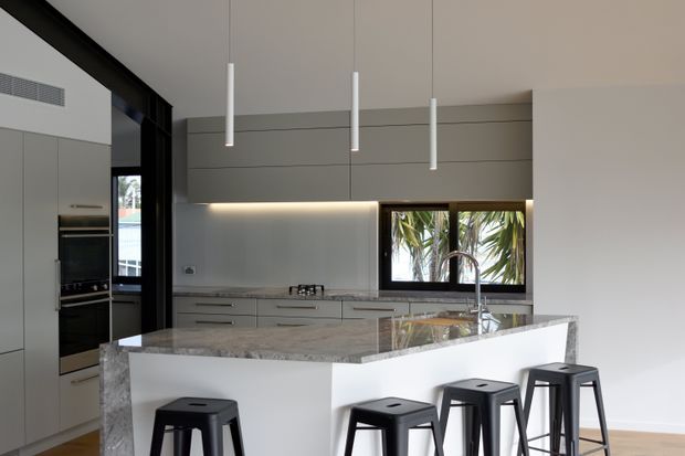 Contemporary kitchen with a large white waterfall island, polished stone benchtop, integrated light-grey cabinetry, dark-framed glazing, and three slim white pendant lights. Black bar stools provide contrast, while tropical foliage is visible through the window.