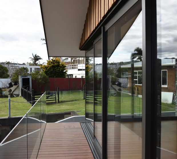 Contemporary residential exterior view from a raised timber deck, featuring extensive dark-framed floor-to-ceiling glazing, a projecting roof overhang, timber vertical screening above the windows, and frameless glass balustrades. The deck overlooks a landscaped suburban garden and neighbouring homes.