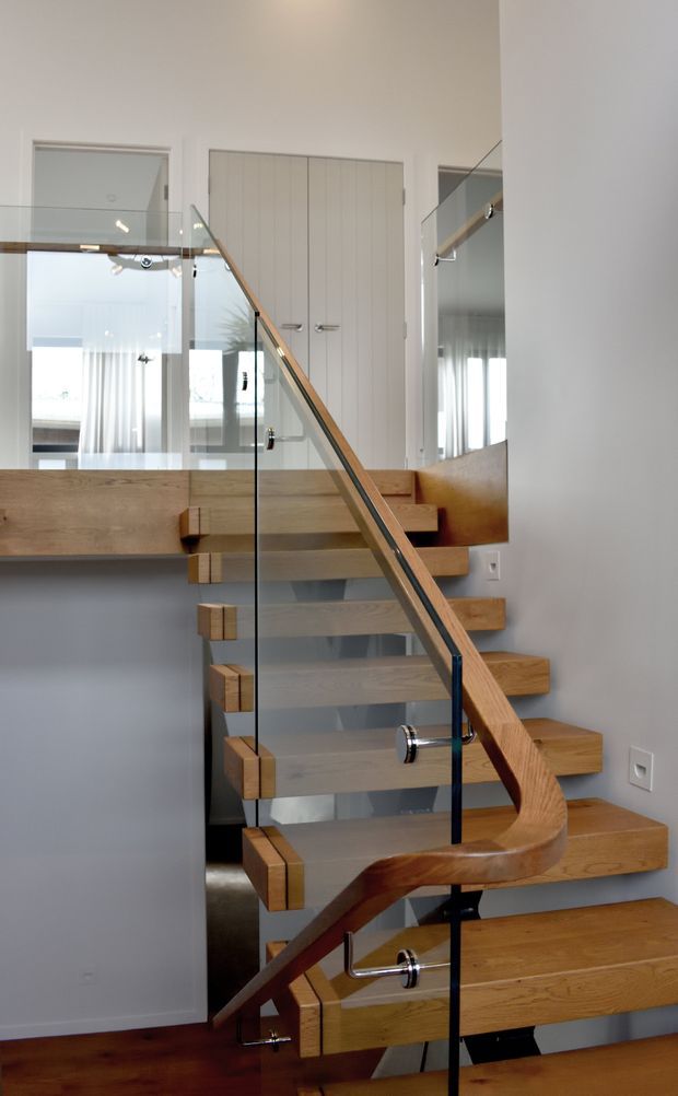 Interior view of a contemporary staircase with floating timber treads, a sculptural curved timber handrail, and clear glass balustrades supported by dark metal fittings. The stair rises through a bright hallway or landing, with white walls, mirrored surfaces, and white paneled doors visible around it.