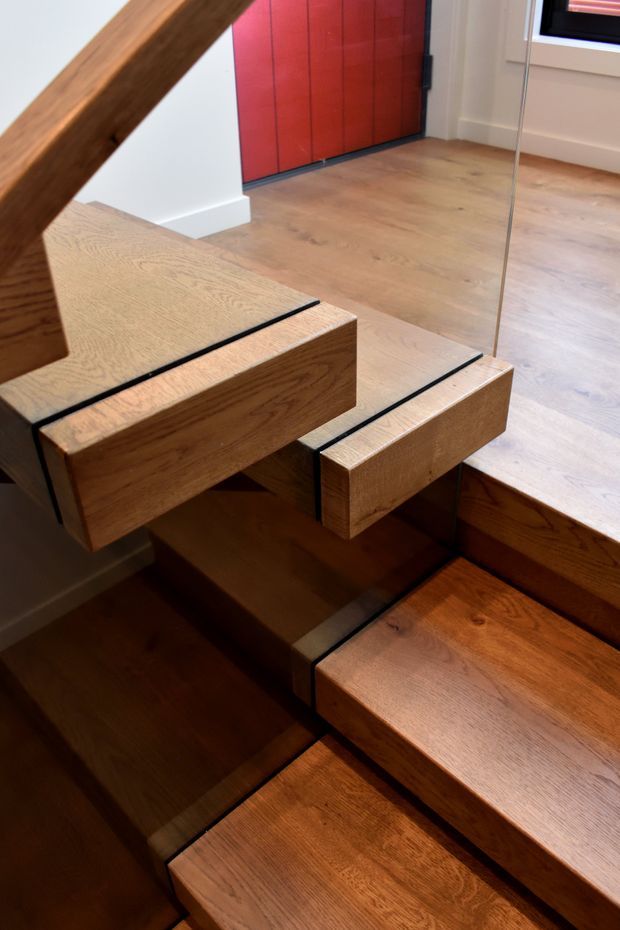 Close-up interior view of a modern timber staircase with thick oak-toned treads and integrated stair stringers, accented by slim black reveals. A frameless glass balustrade runs alongside the stairs. The landing has matching timber flooring, a vivid red flush door, white skirting, and a black-framed window.