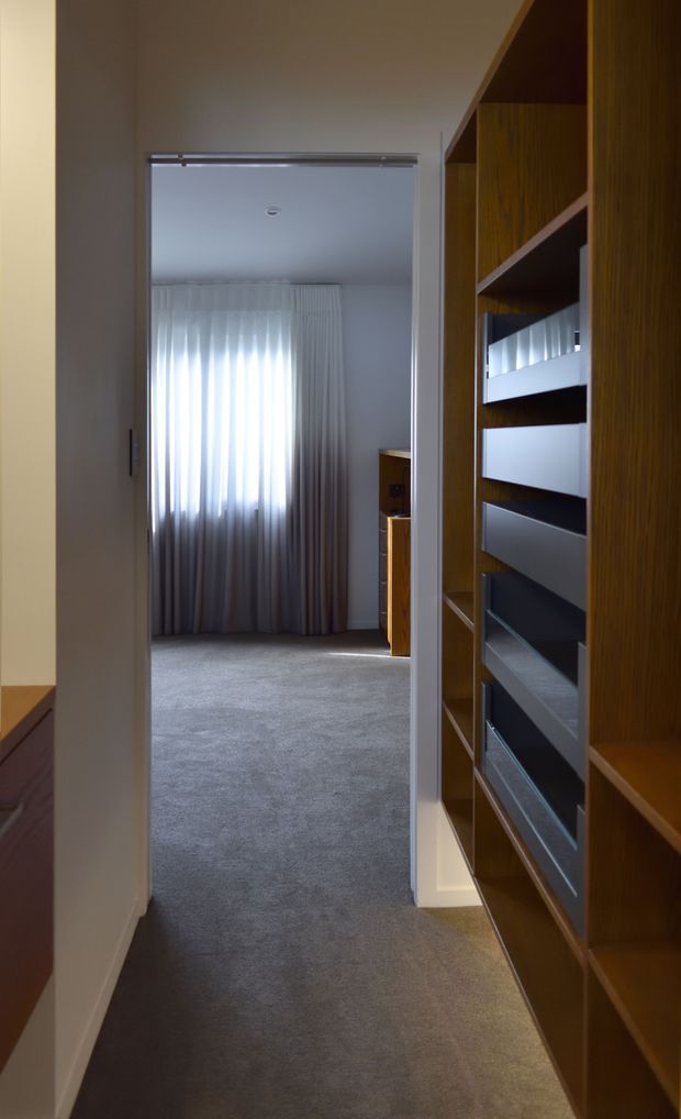 A view down a narrow hallway toward a softly lit carpeted room with full-height sheer curtains. The hallway features extensive built-in timber shelving and drawers on the right, with neutral painted walls and grey carpeting. The room beyond contains additional timber joinery and a small glimpse of furniture.