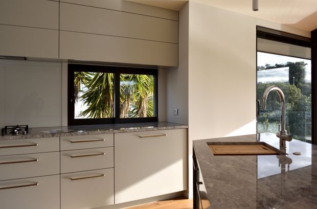 Contemporary kitchen with extensive matte taupe cabinetry, long horizontal handles, a dark-framed window, and a polished stone benchtop. A central island with an undermount sink, curved chrome tap, and timber chopping board faces full-height glazed doors opening to a leafy exterior. Warm sunlight creates a refined, understated atmosphere.