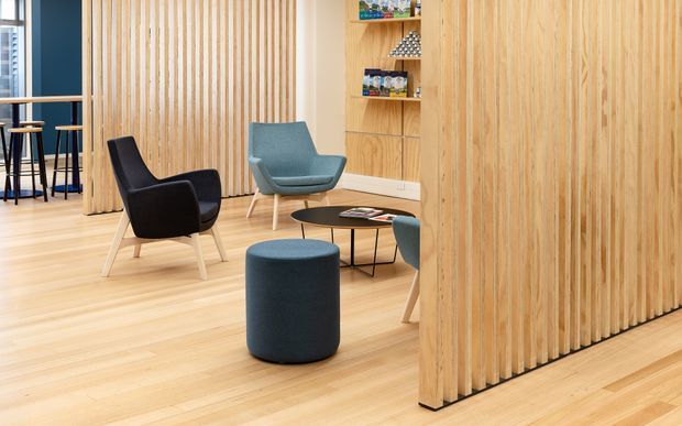 A contemporary commercial lounge or waiting area featuring pale timber flooring and extensive vertical timber slat wall screens. A black lounge chair, muted blue lounge chair, round blue ottoman, and low round coffee table form a seating arrangement. Open shelving displays small items, while a bar-height seating area is visible beyond the slatted partition.
